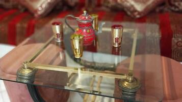 Una mesa con cubierta de vidrio y patas de metal ornamentadas, una tetera roja y dos jarrones pequeños sobre un sofá estampado.
