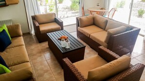 Sala de estar con muebles de mimbre, una mesa de centro y vistas a un jardín a través de amplios ventanales.