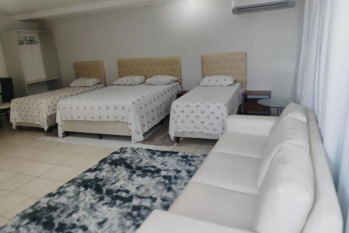 A hotel room with three single beds, a white sofa, a black and white rug, and an air conditioning unit on the wall.