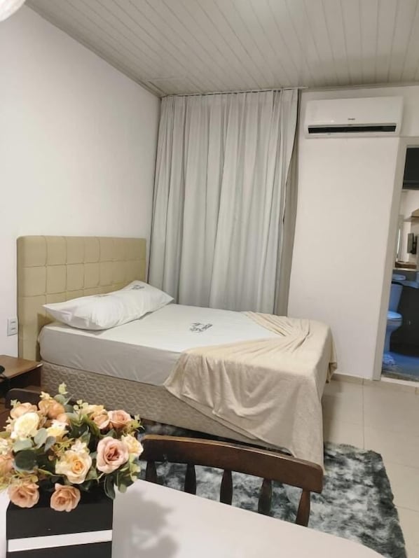 A small, neatly arranged bedroom with a single bed, a nightstand, a chair, and a table with a vase of flowers. - Hotel veneza (Itabuna)