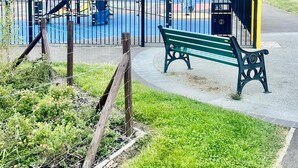 A playground with a slide and climbing structures, surrounded by a fence, with a bench and a street in the background.