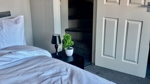 A neatly made bed with white bedding, a small bedside table with a black lamp and a potted plant, and a closed door in the background.
