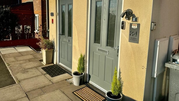 A two-story house with a grey door and a security camera. There are two potted plants on the ground floor.