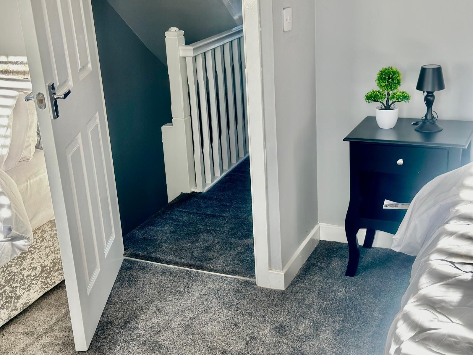 A hallway with a carpeted floor, a white door, a staircase, a dark-colored dresser with a lamp, and a small potted plant.