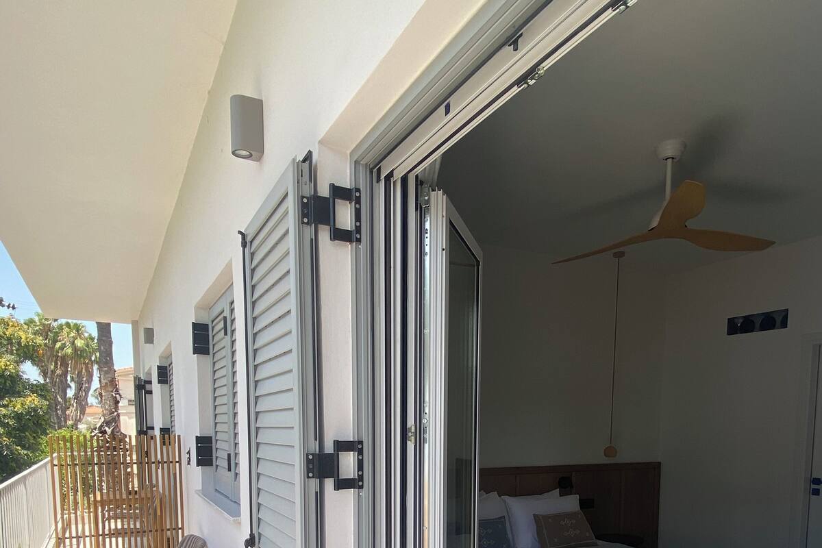 Un balcon avec une table et des chaises, une porte-fenêtre coulissante donnant sur une chambre à coucher équipée d’un lit et d’un ventilateur de plafond.