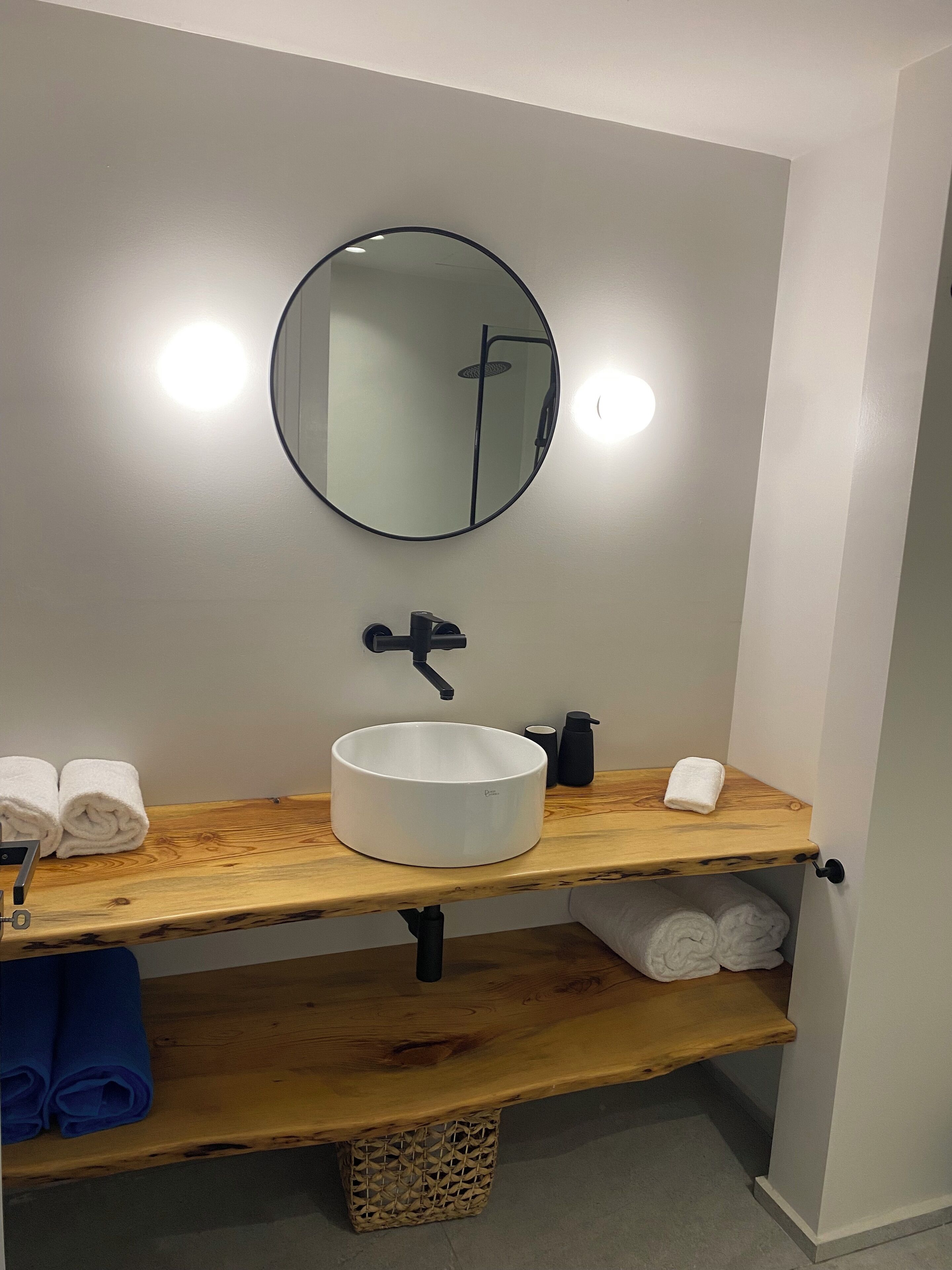 A bathroom with a round mirror, a white sink, and wooden shelves holding towels.
