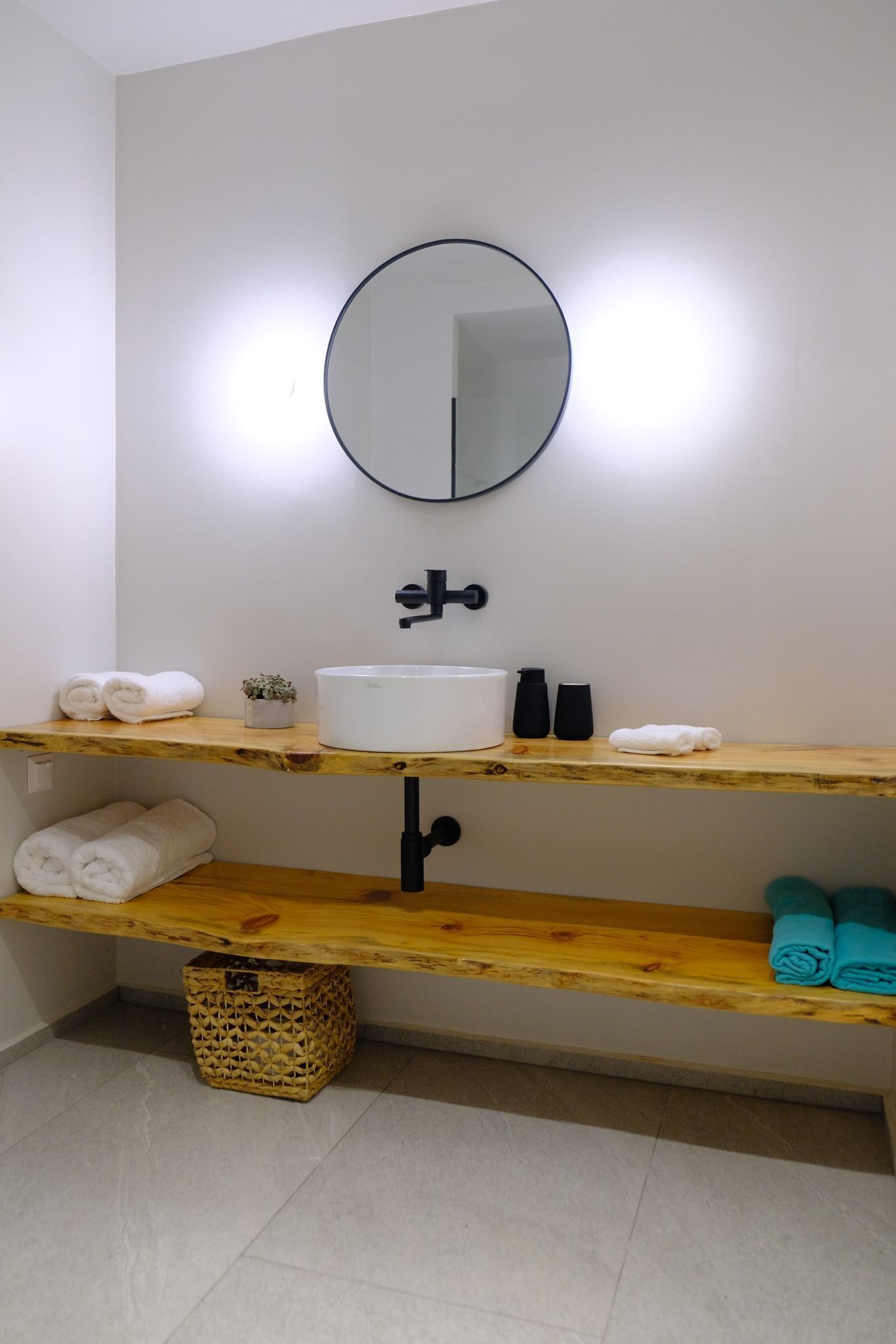 Een badkamer met een wit wastafelmeubel, een ronde spiegel en houten planken met handdoeken en toiletartikelen.