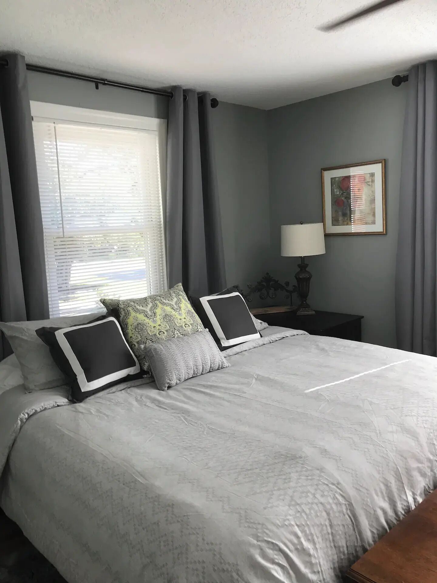 A neatly made bed with a grey and white patterned bedspread, two black pillows with white trim, and a grey throw pillow. There is a window with blinds, a framed picture on the wall, and a small table with a lamp beside the bed.