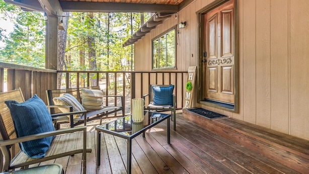 A wooden porch with a glass table and chairs, a wooden door with a 'HOME' sign, and a view of trees.