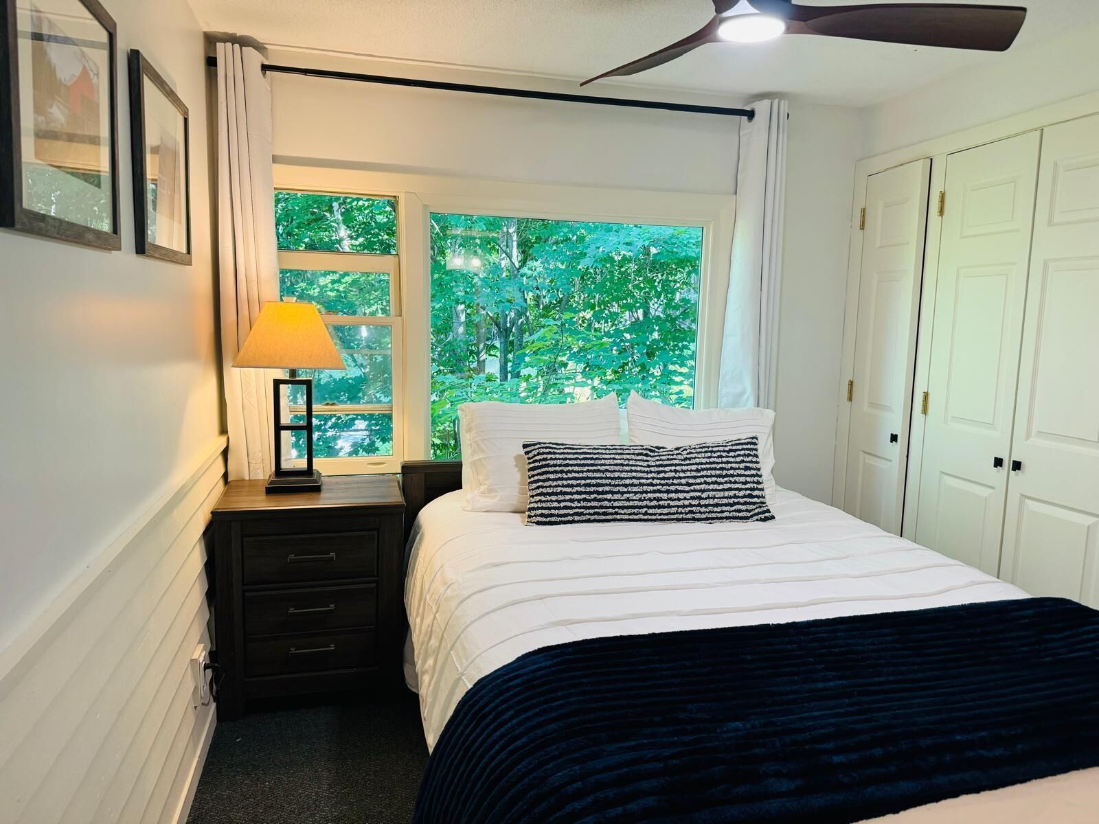 A bedroom with a bed, a nightstand, a lamp, a window with a view of trees, and a ceiling fan.