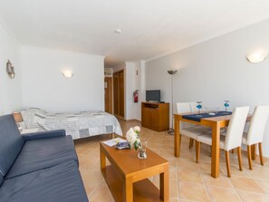 A modern living room with a sofa, a wooden coffee table, a dining table with chairs, a TV on a wooden cabinet, and a patterned bedspread. - Quinta Velha Village Studio with Pool (Cabanas de Tavira)
