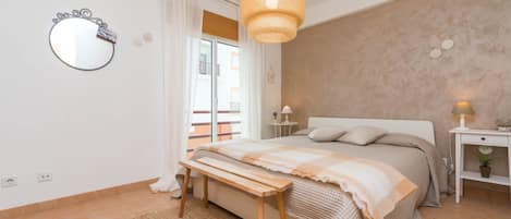 A bedroom with a bed, bedside tables, a hanging light fixture, a mirror, and a view of the outside.