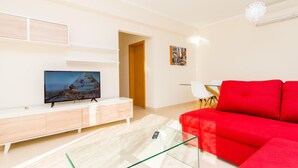 Ein modernes Wohnzimmer mit einem Flachbildfernseher, einem roten Sofa, einem Glastisch und einem Essbereich mit einem weißen Tisch und Stühlen.