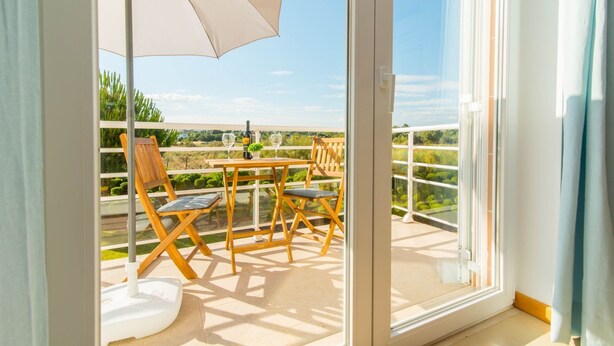 Un balcón con mesa y sillas, una sombrilla blanca y vistas a zonas verdes.