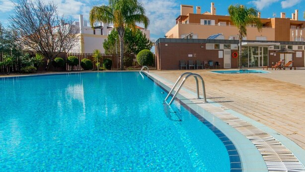 Piscina azul cristalina com escada de metal, rodeada por um deck de madeira e palmeiras.