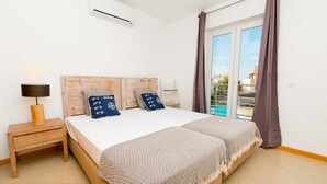 Quarto com cama, mesa de cabeceira, luminária e vista para o exterior através de uma porta de correr de vidro.