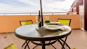 A round glass table with a yellow umbrella, two chairs, a bottle of wine, and a small plant on a balcony with a view of the sky and a building.