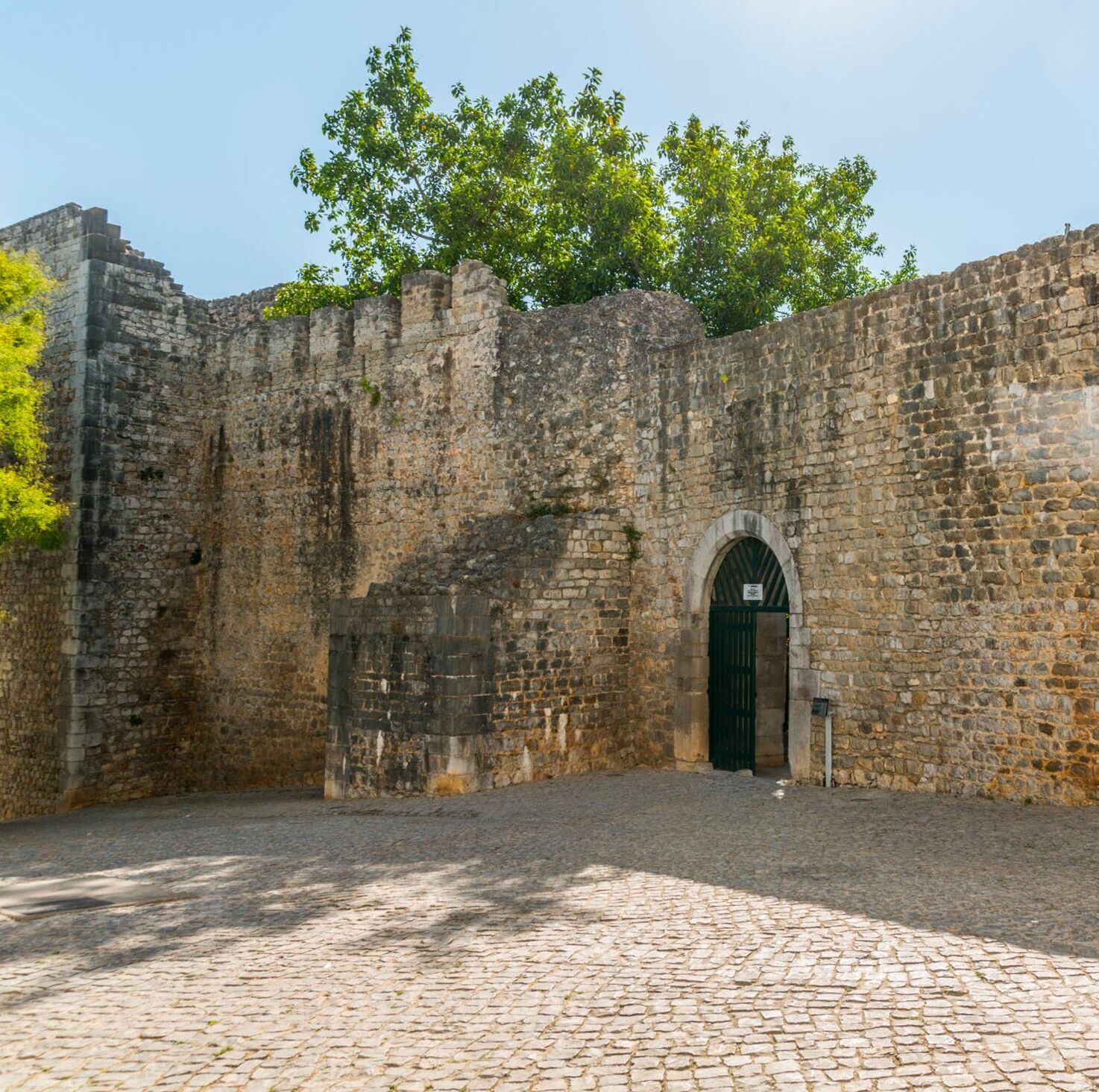 A historic stone wall with a green arched entrance, cobblestone ground, and trees in the background.