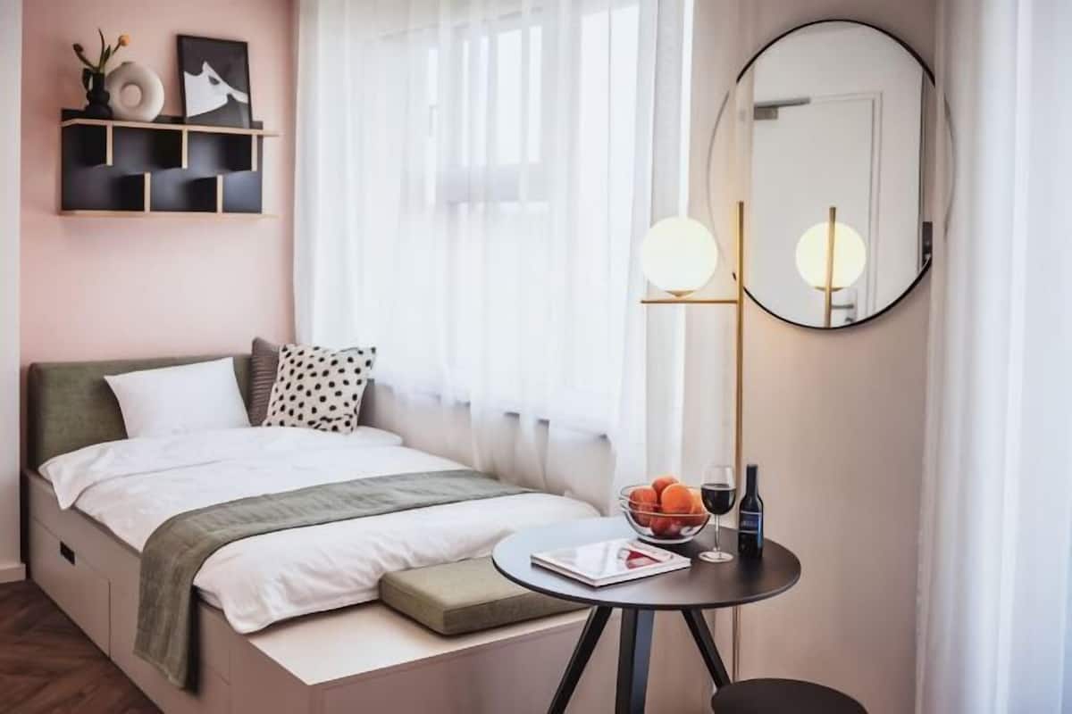 Ein Schlafzimmer mit Bett, Nachttisch, Spiegel und einem Fenster mit durchsichtigen Vorhängen.
