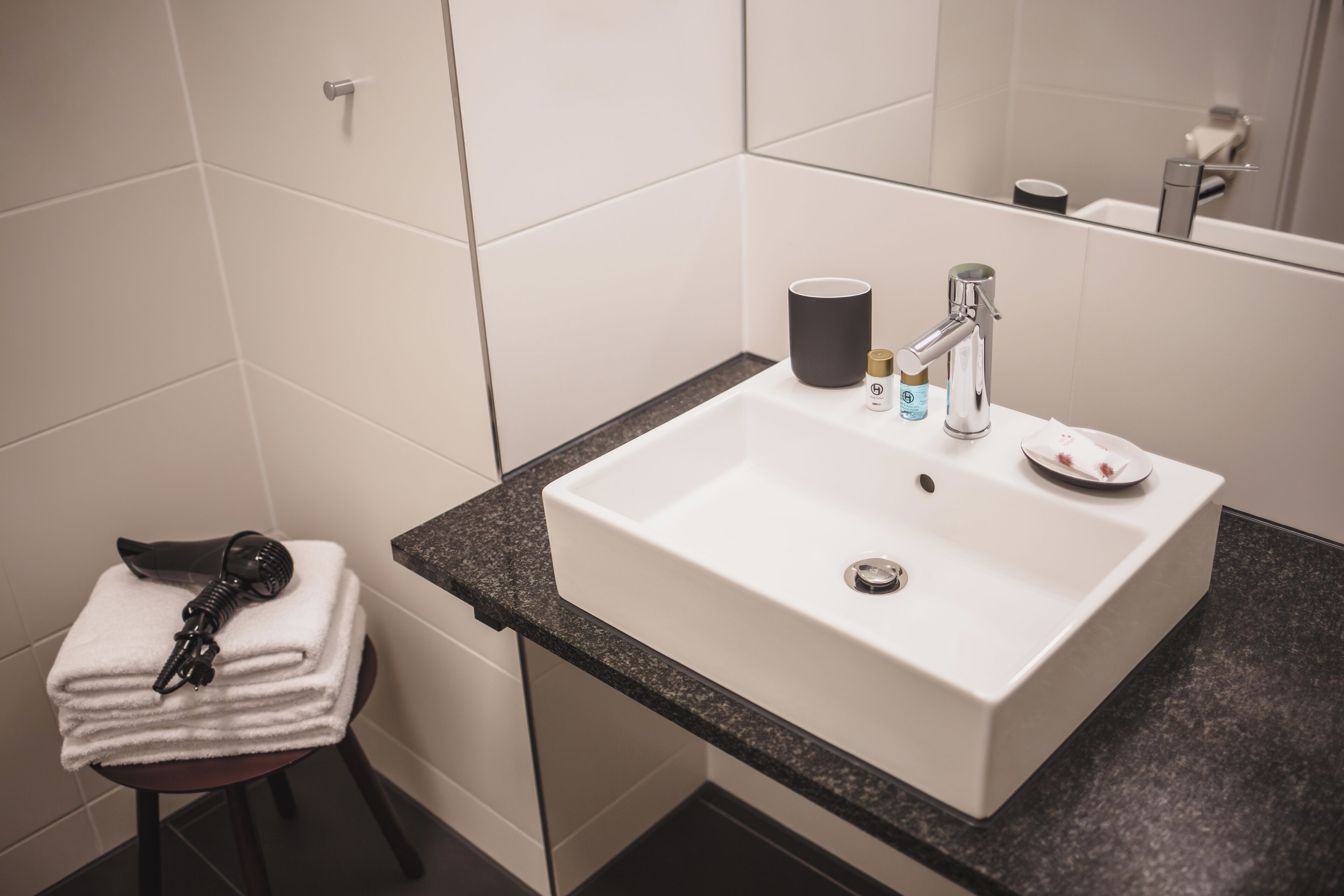 Un baño con un lavamanos blanco, encimera oscura y un espejo que refleja un inodoro y un perchero.