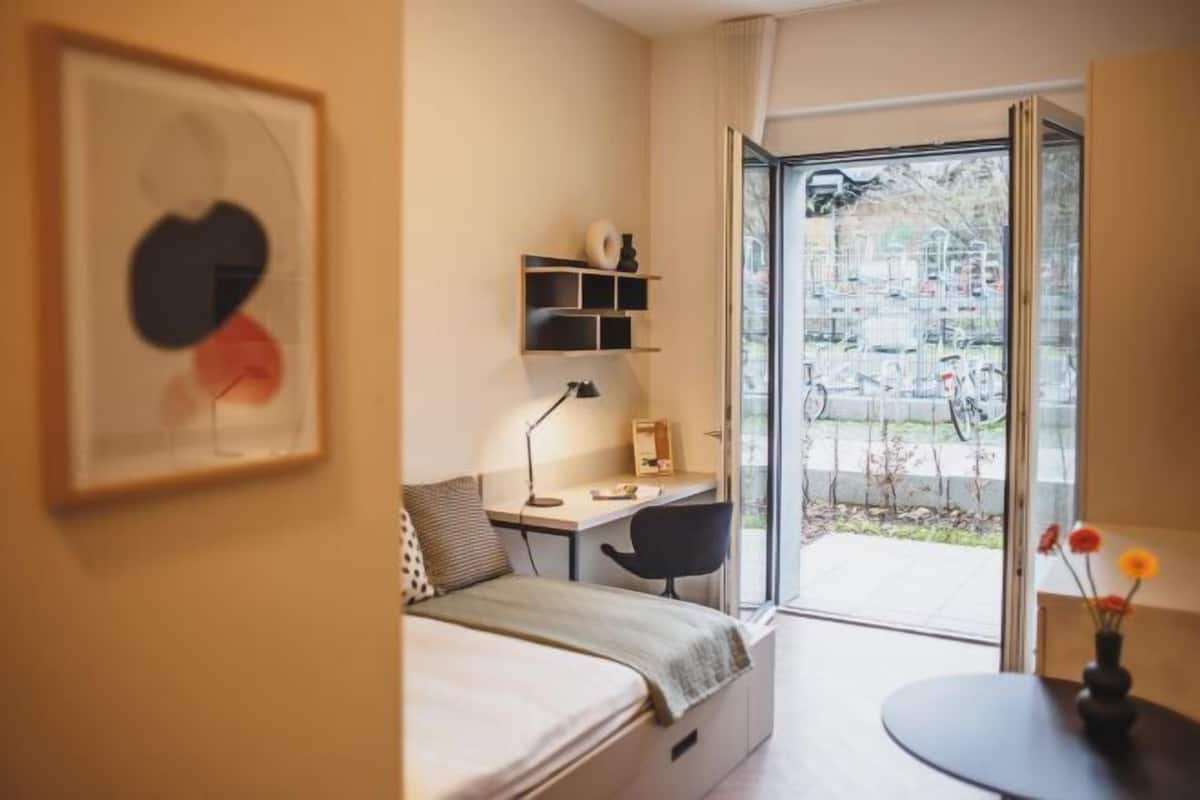 Ein Schlafzimmer mit Bett, Schreibtisch, Stuhl und Blick durch eine Glastür.