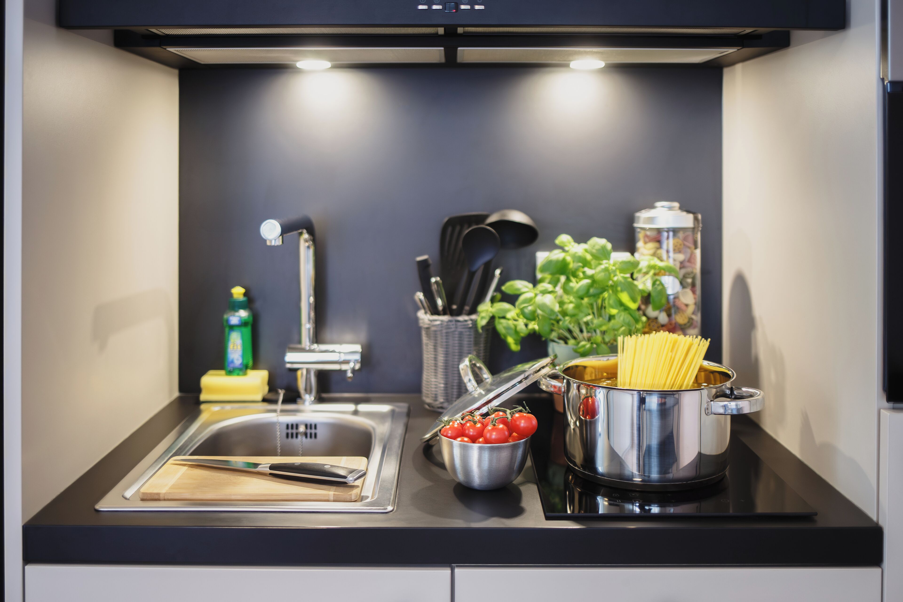 A modern kitchen with a sink, cutting board, bowl of tomatoes, and a pot of spaghetti.