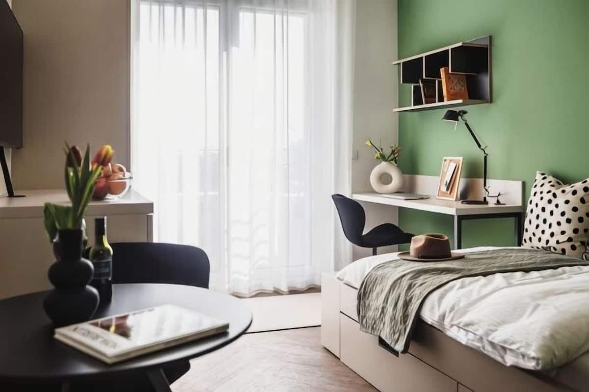 Ein Schlafzimmer mit Bett, Schreibtisch, Stuhl, einem Fenster mit Vorhängen und einem Regal mit Büchern und einer Vase.