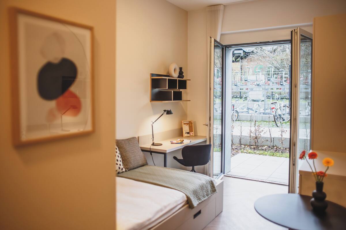 Ein modernes Schlafzimmer mit Bett, Schreibtisch, Stuhl und Blick ins Freie.