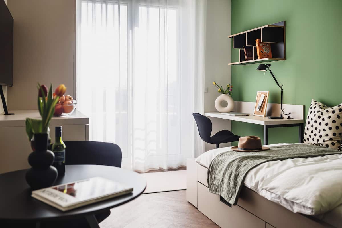 Ein modernes Schlafzimmer mit einem Bett, einem Schreibtisch und einem Sessel. Ein Fenster mit durchsichtigen Vorhängen, eine Vase mit Blumen und ein Bücherregal sind ebenfalls vorhanden.