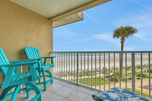 A balcony with turquoise chairs and a patterned cushion, overlooking a beach with a palm tree and a clear sky.
