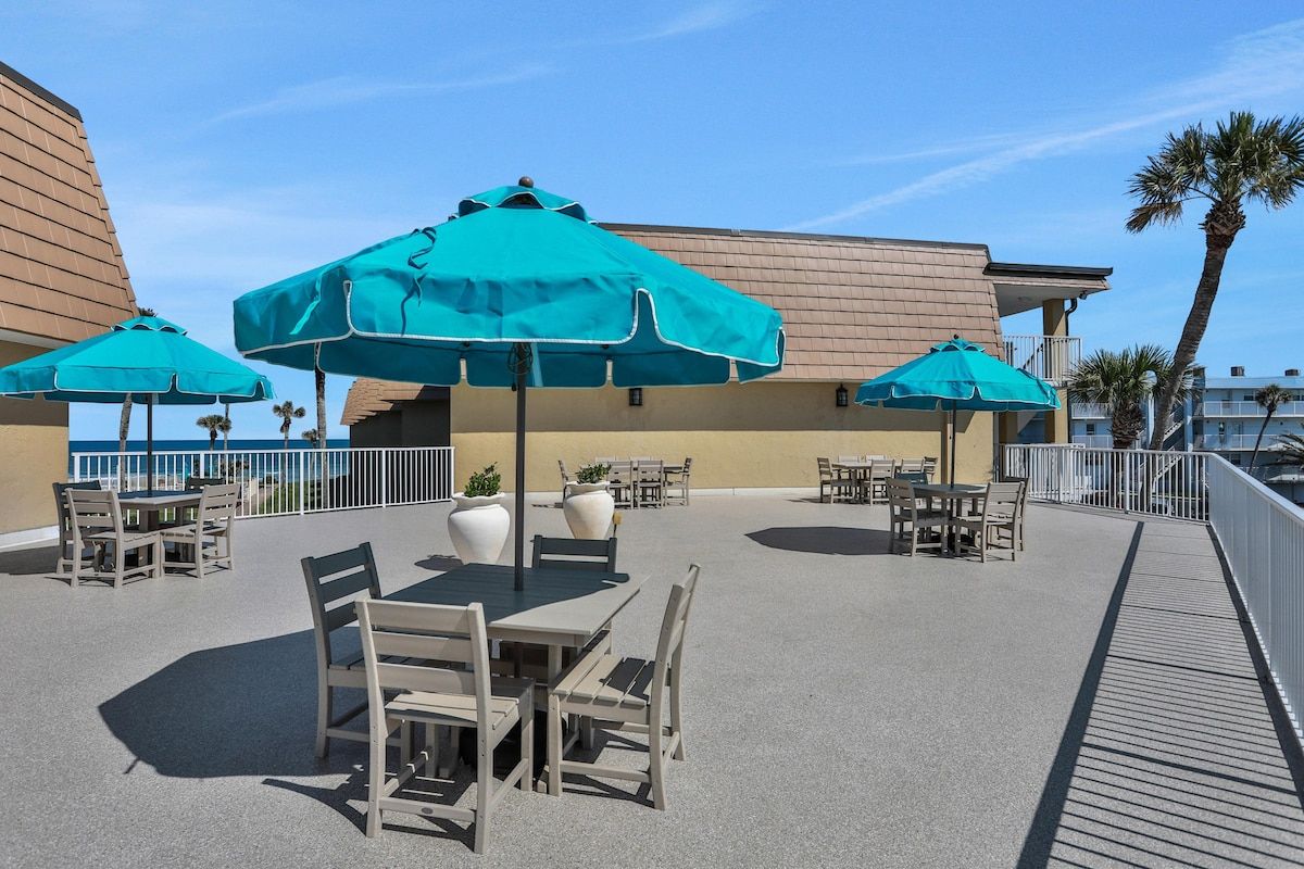 An outdoor patio area with tables and chairs, shaded by large umbrellas, and a view of the ocean.