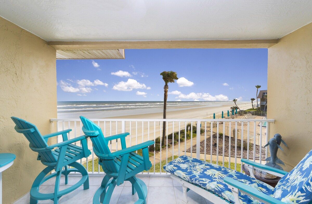 A balcony with turquoise chairs and a sofa, overlooking a beach with palm trees and a clear sky.