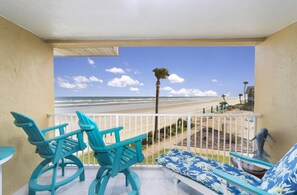 A balcony with turquoise chairs and a sofa, overlooking a beach with palm trees and a clear sky.