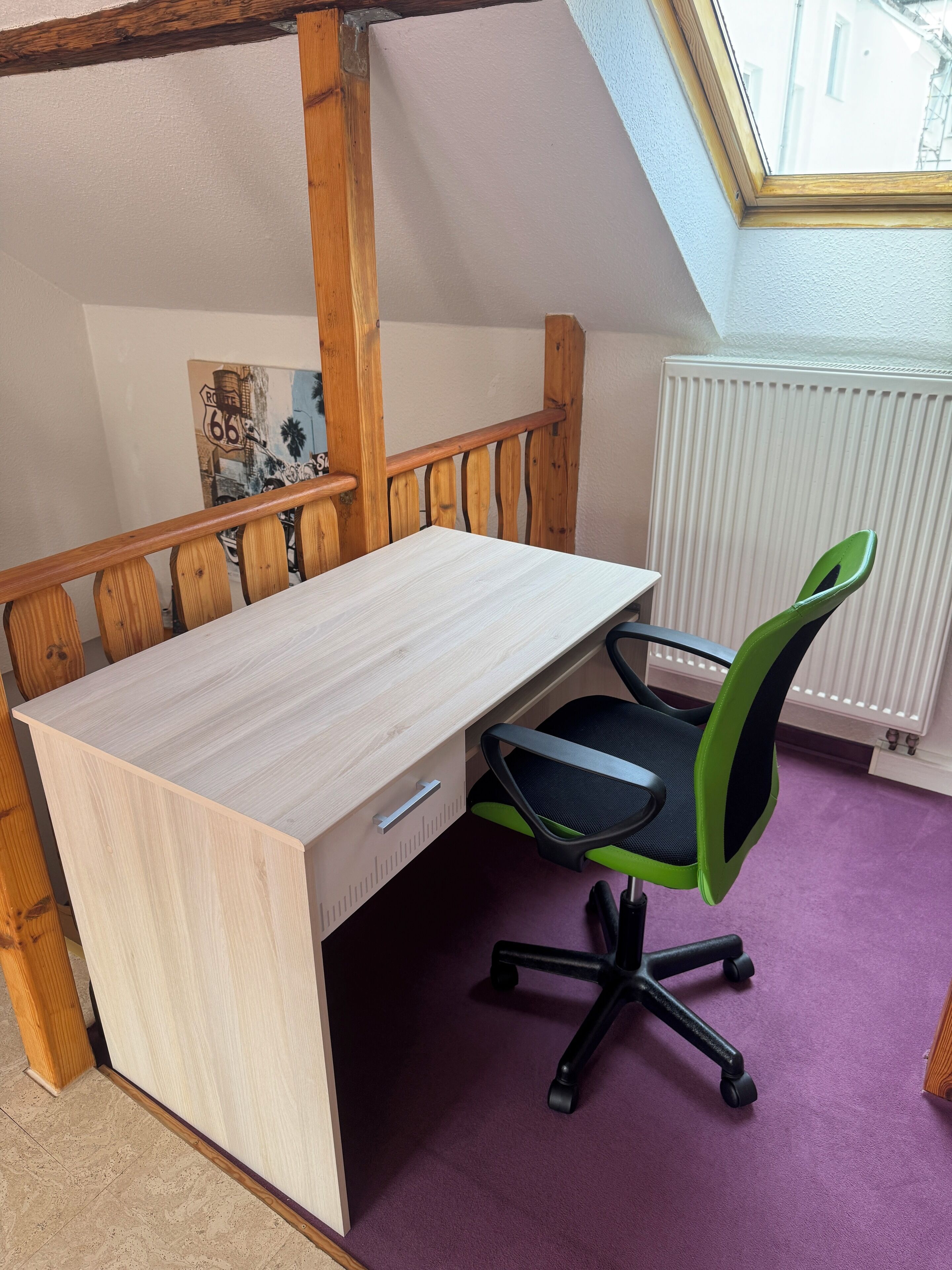 A room with a sloped ceiling, a wooden desk with a drawer, a green and black office chair, and a purple carpet.