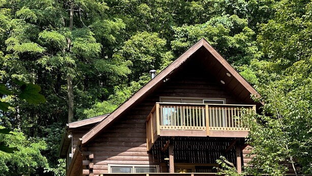 A two-story log cabin with a balcony on the upper floor, surrounded by dense forest. - Charming 3-bdrms & loft cabin, with WiFi, mins to Elk Mountain by Lake Idlewild (Clifford)
