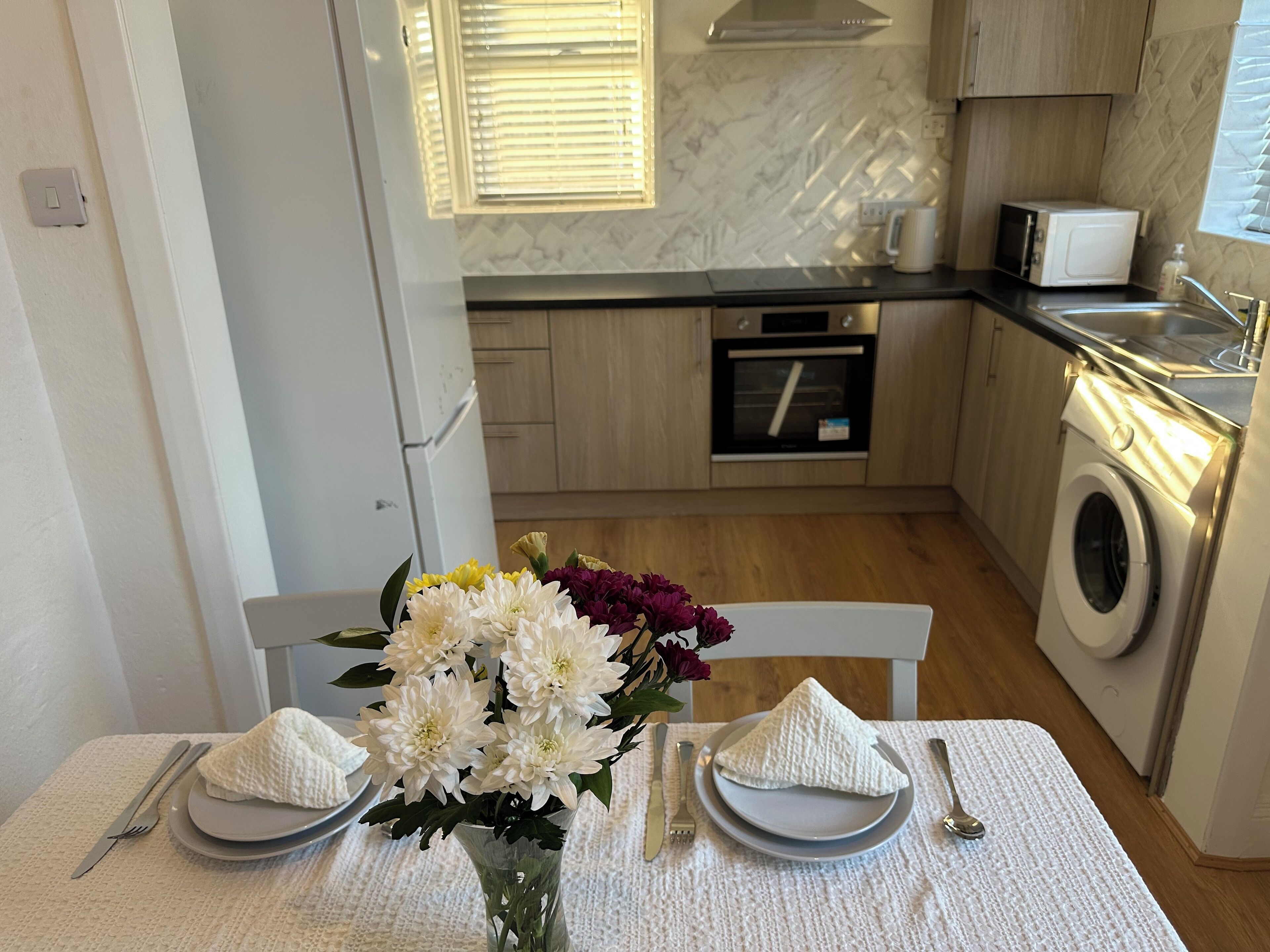 Un comedor con una mesa dispuesta para dos personas, un jarrón de flores y una cocina al fondo.
