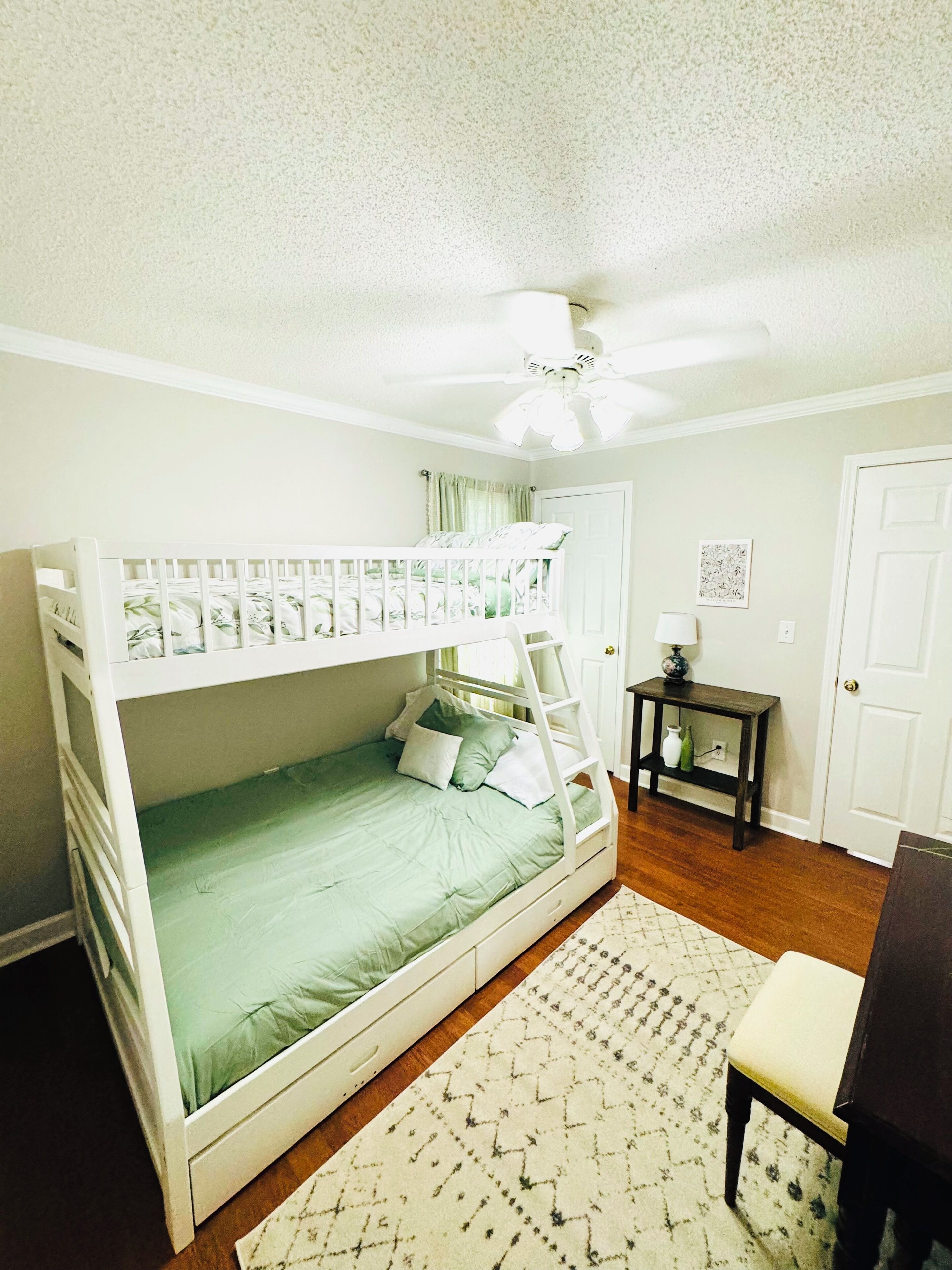 A bunk bed with a ladder, a small table with a lamp, a door, and a patterned rug.