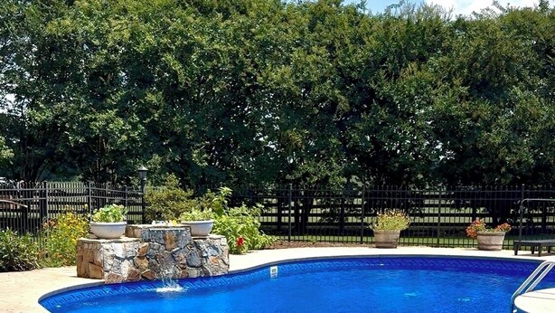 Une piscine bordée de pierres, entourée d’une clôture et de verdure.