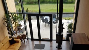 A spacious room with a large window offering a view of a green landscape. There is a sofa, a dining table with chairs, and a fireplace. - Modern chalet - relaxing stay with private spa, calm, nature and comfort (RUYNES EN MARGERIDE)