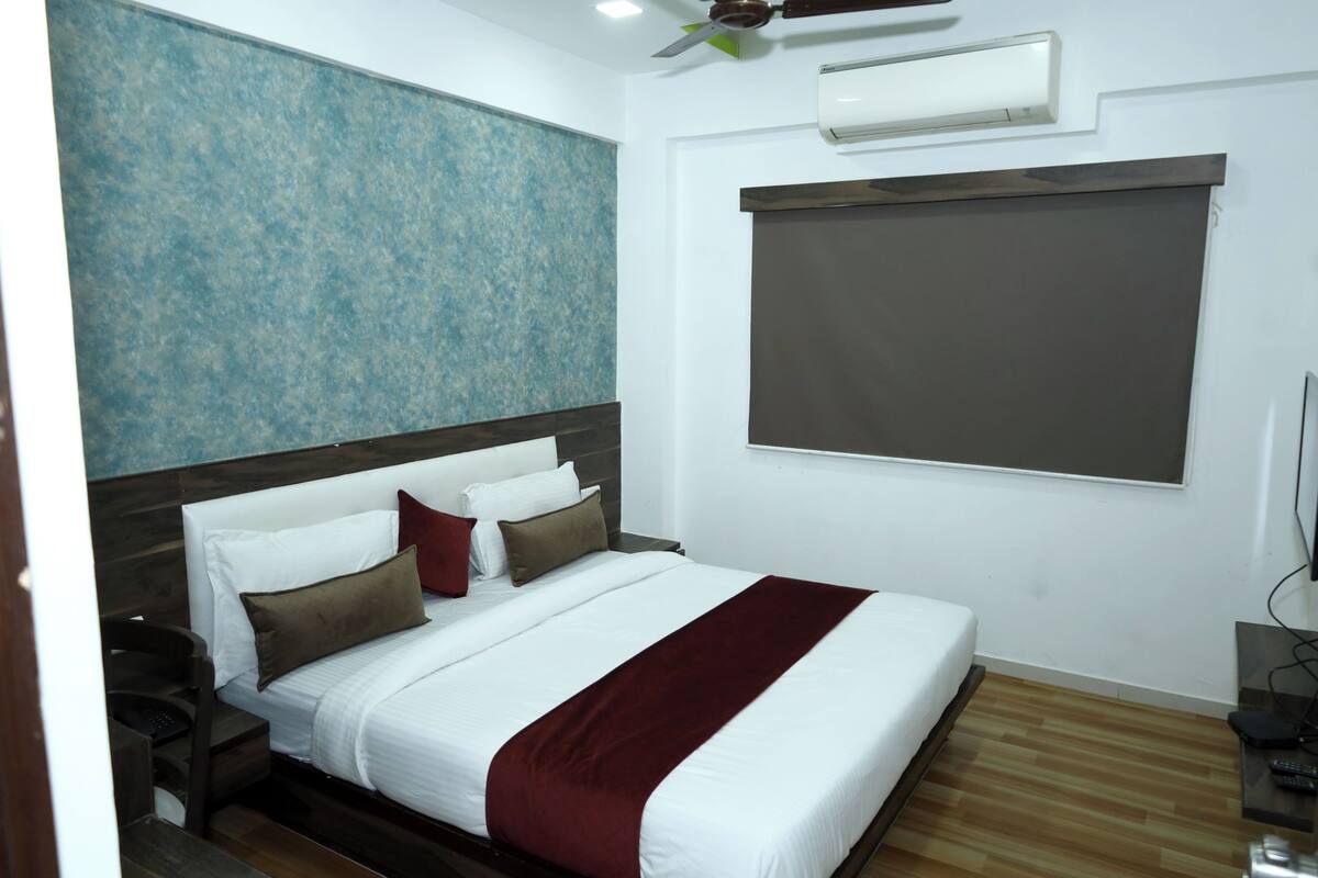 A hotel room with a large bed, a ceiling fan, an air conditioning unit, a wooden floor, and a large window with a blind.