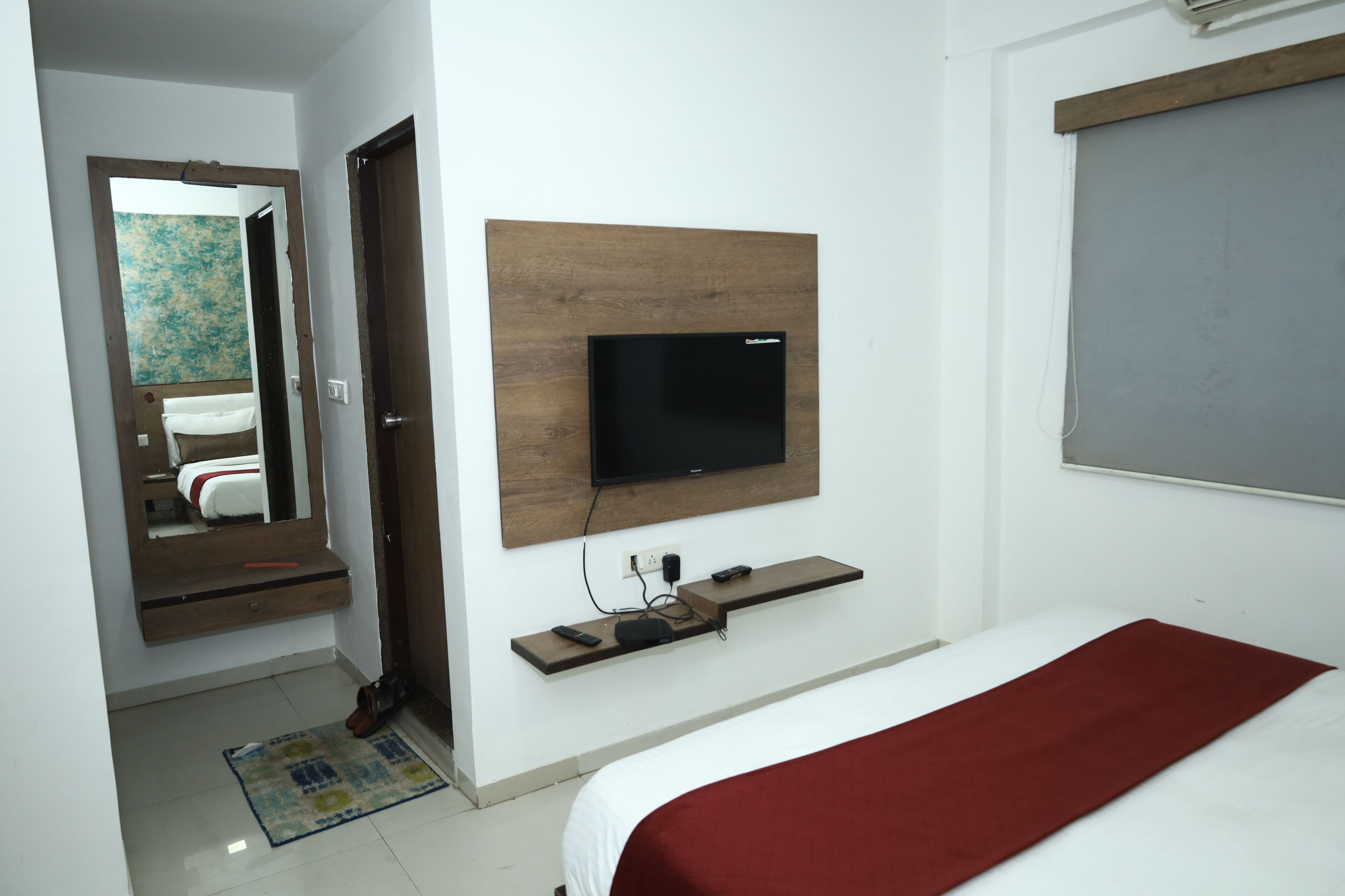 A hotel room with a bed, a TV mounted on a wooden panel, a mirror, and a small shelf with a remote control.