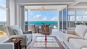 A spacious room with a large glass door leading to a balcony with a view of the ocean. There are wicker chairs and a sofa, a glass coffee table, and a ceiling fan.