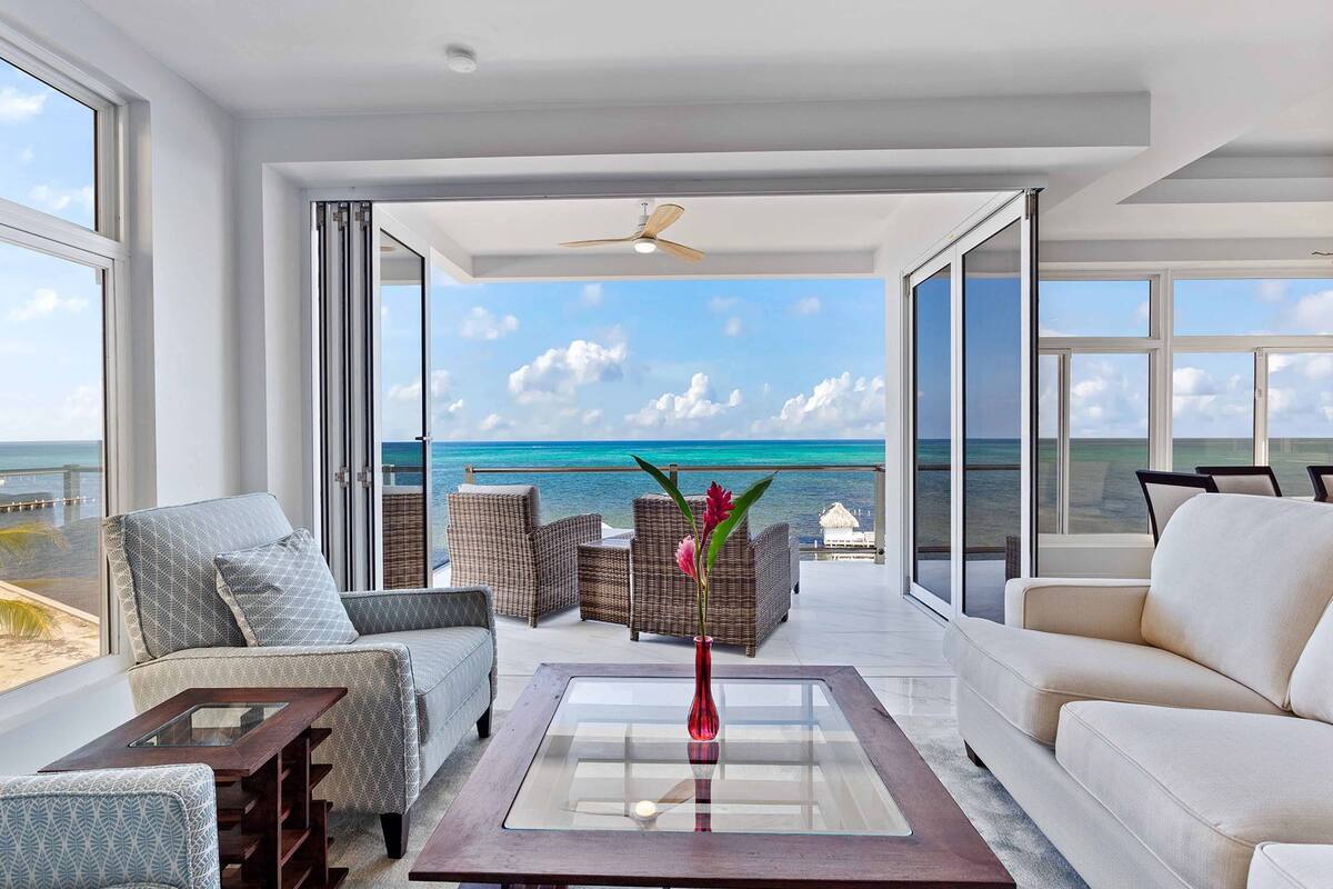 Ruime kamer met een grote glazen deur die leidt naar een balkon met uitzicht op de oceaan. Er staan rieten stoelen en een bank, een glazen salontafel en een plafondventilator.