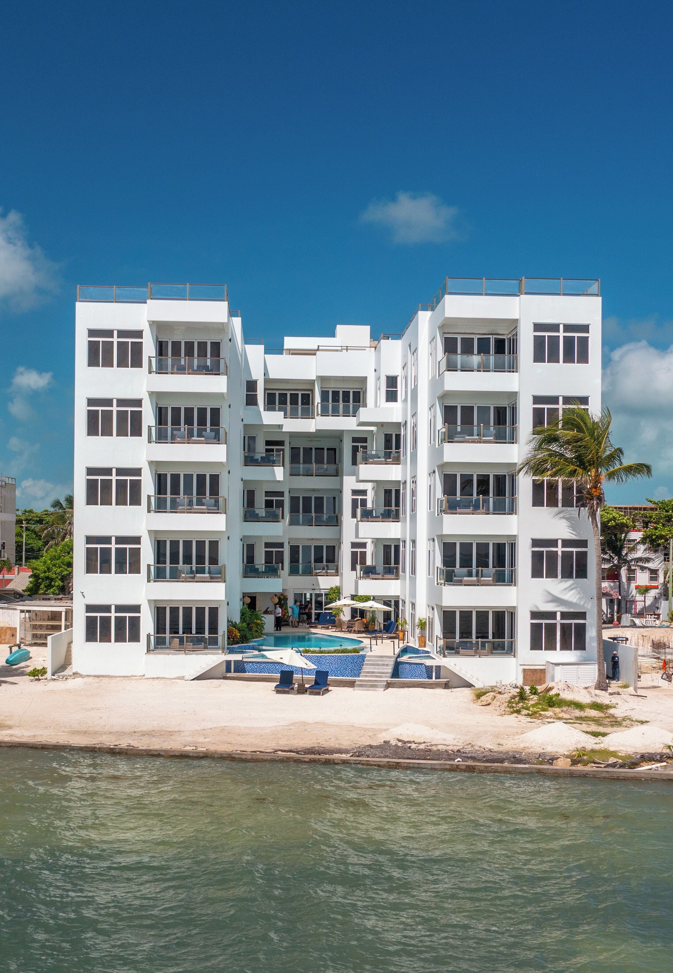 A white multi-story hotel with balconies, a pool, and a beach view.