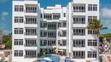 A white multi-story hotel with balconies, a pool, and a beach view.