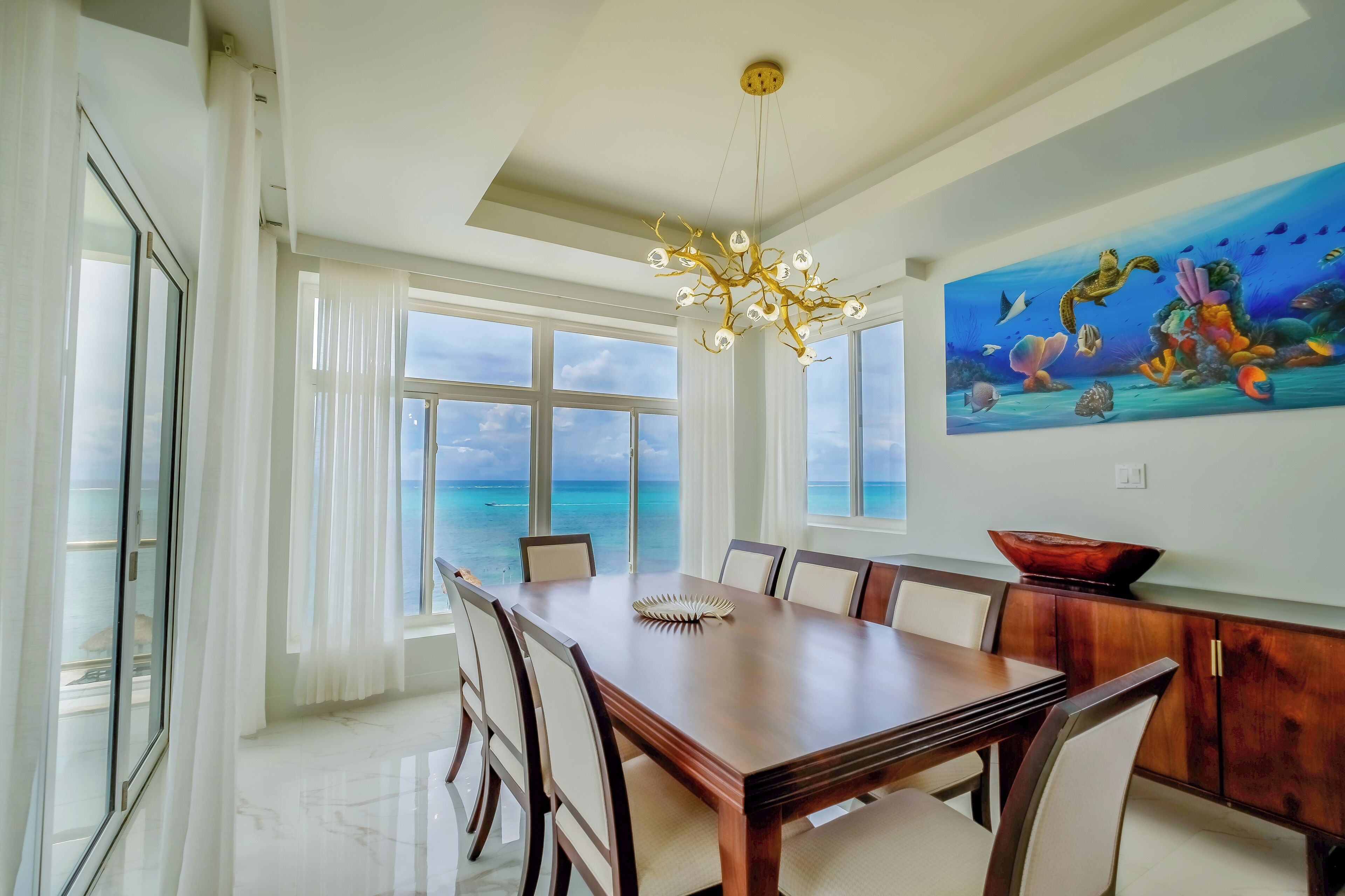 A dining area with a large table, chairs, a chandelier, a painting of an underwater scene, and a view of the ocean through large windows.