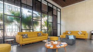 A modern living room with two yellow sofas, a grey ottoman, and a wooden coffee table with magazines. Large windows allow natural light and views of palm trees. - Your cofortable Oasis in Playa del Carmen (Playa del Carmen)