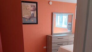 A bedroom with a wooden floor, a dresser, a bed, and a window with blinds.