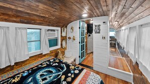 A room with wooden ceilings, a bed with a colorful blanket, a window with curtains, and a door leading to another room.
