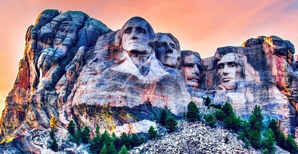 A large rock formation with carved faces, surrounded by trees and a clear sky.